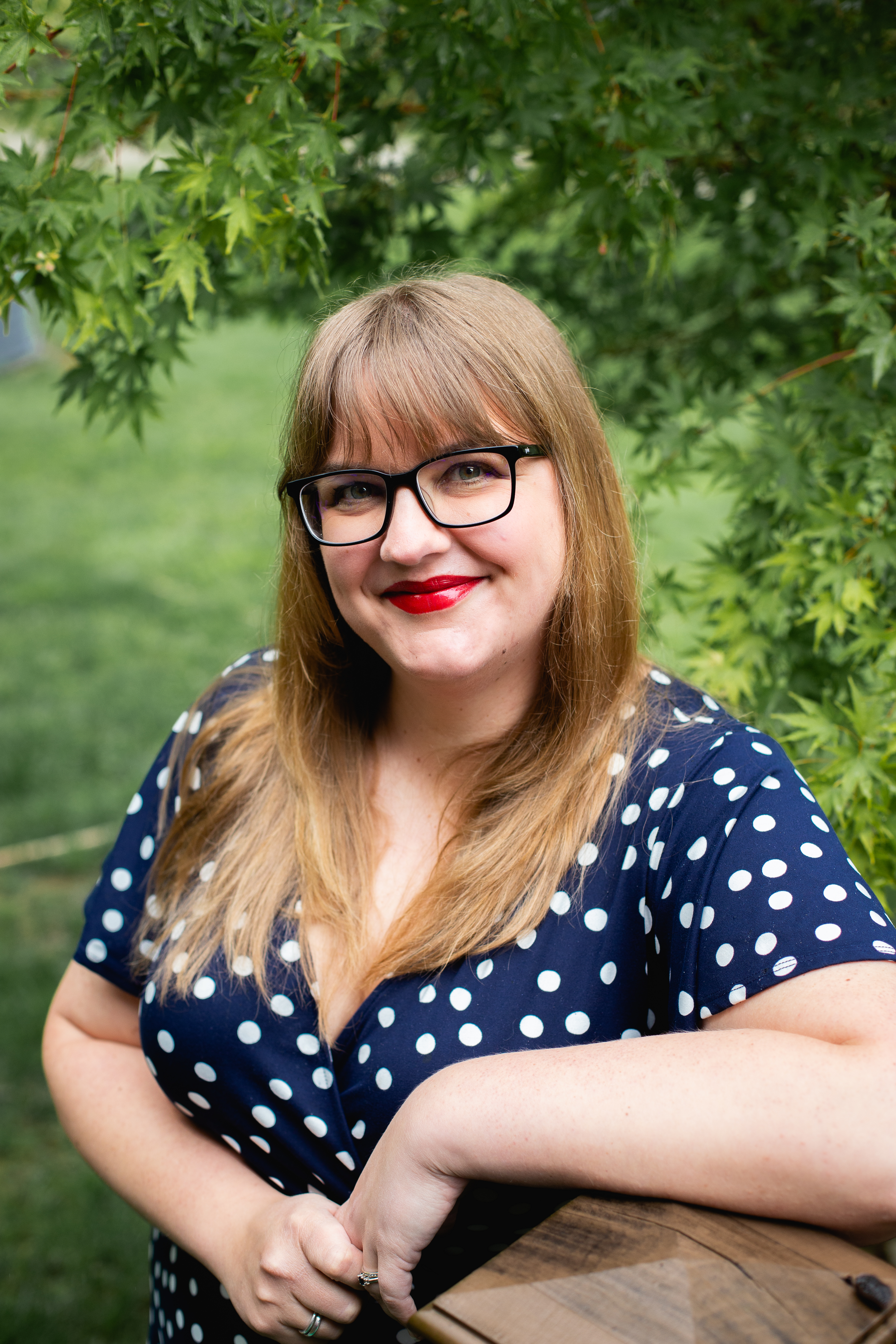 Lindsay Rochester smiling outdoors beneath green maple leaves, wearing glasses and a navy polka-dot dress